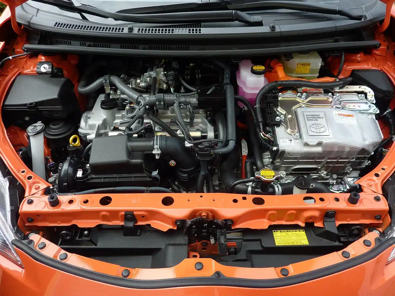 Car battery under an open hood with a hand pointing to the case and terminals