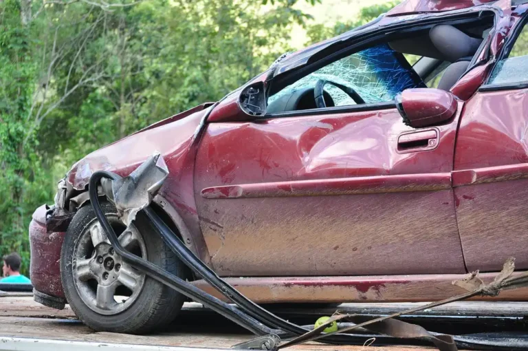 Parent holding a scuffed car seat beside a damaged parked car in daylight.