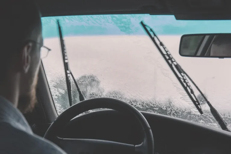 Driver beside a car holding a windshield wiper arm and replacement blade near the windshield