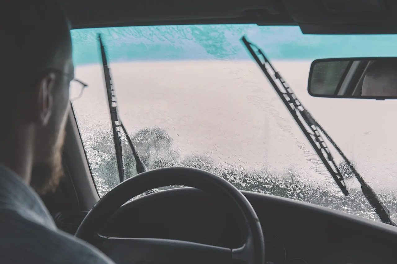 Driver beside a car holding a windshield wiper arm and replacement blade near the windshield